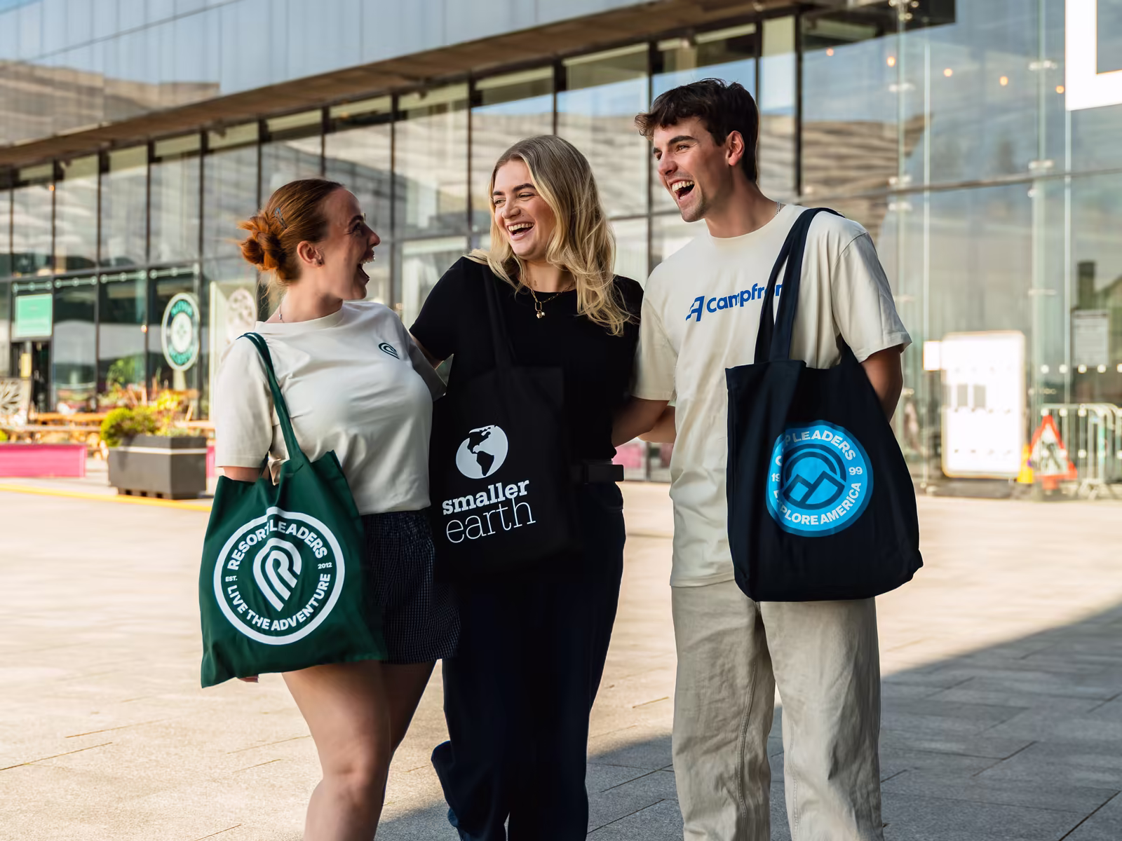 Three colleagues laughing together as they walk through Liverpool, carrying Resort Leaders, Smaller Earth and Camp Leaders tote bags