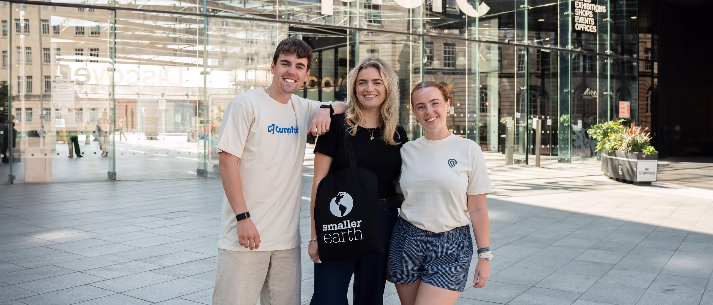 Three Smaller Earth colleagues smiling outside a glass-fronted building in Liverpool, in Campfront and Resort Leaders T-shirts and carrying a Smaller Earth tote