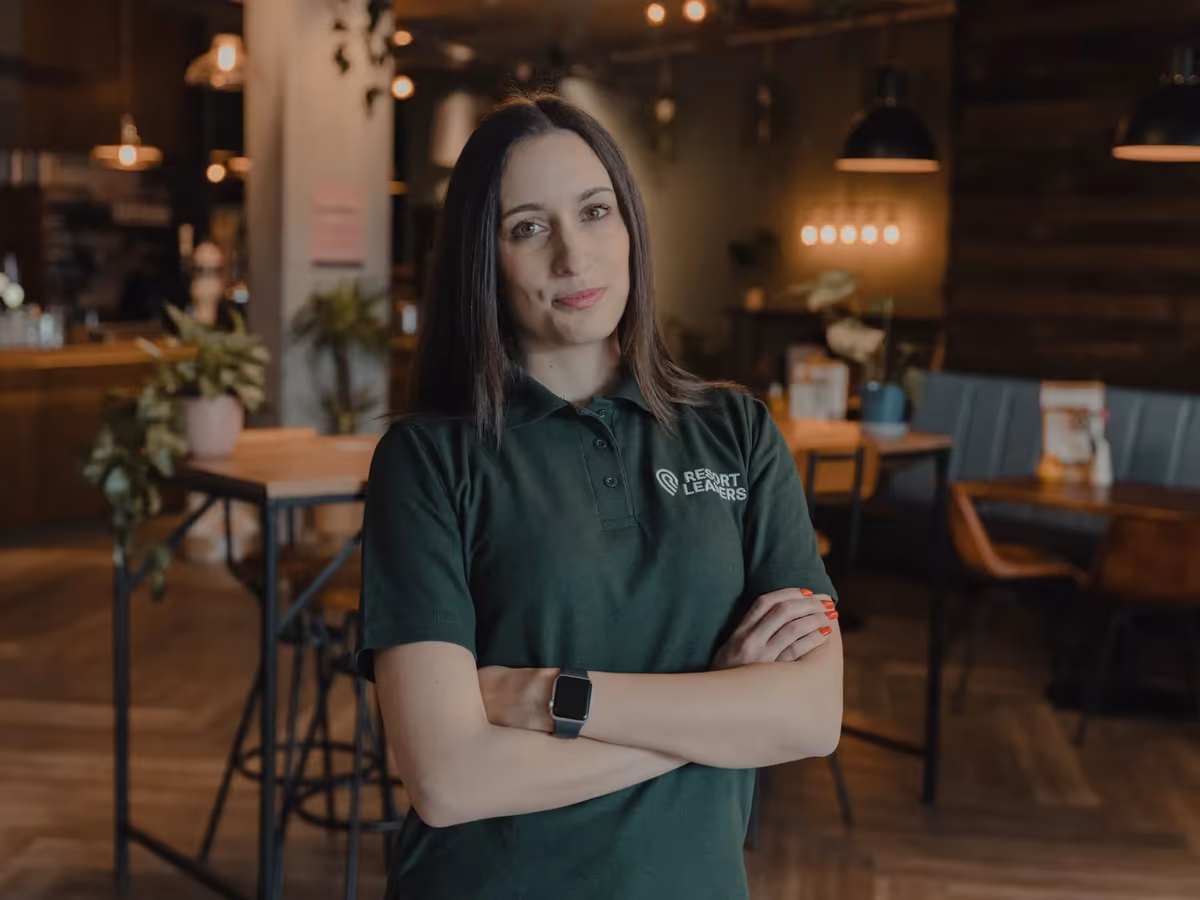 A Resort Leaders staff member in a branded green polo shirt standing with her arms folded in a resort restaurant