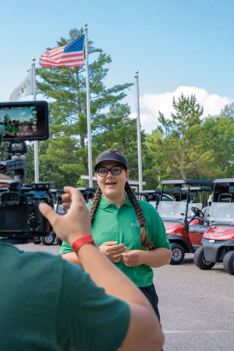 A resort staff member in a green polo shirt being filmed on camera outside, flags and golf carts behind her