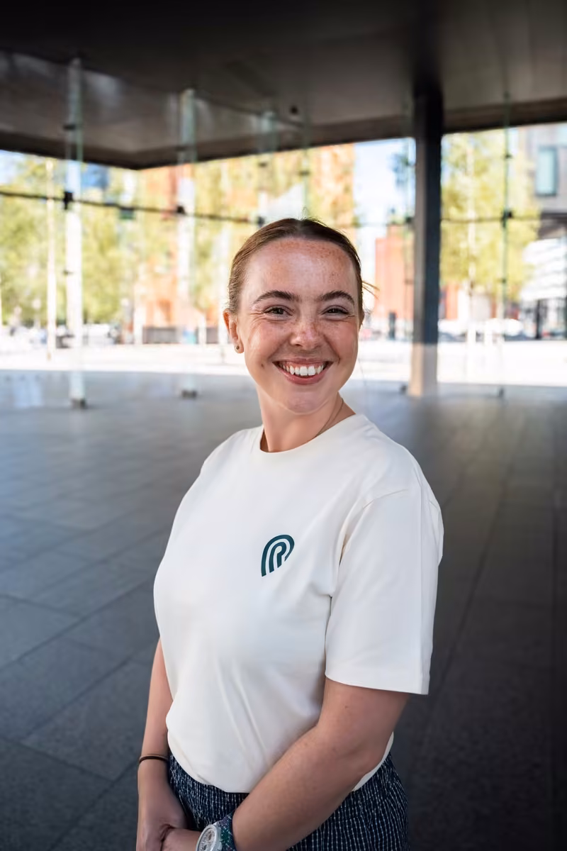 A smiling woman with freckles in a cream Resort Leaders T-shirt, standing under a glass canopy in Liverpool