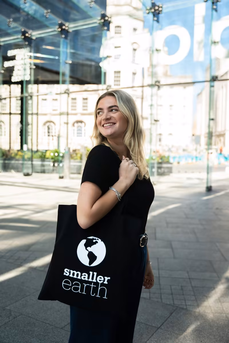 A woman in a black T-shirt glancing back over her shoulder with a Smaller Earth tote bag on her arm