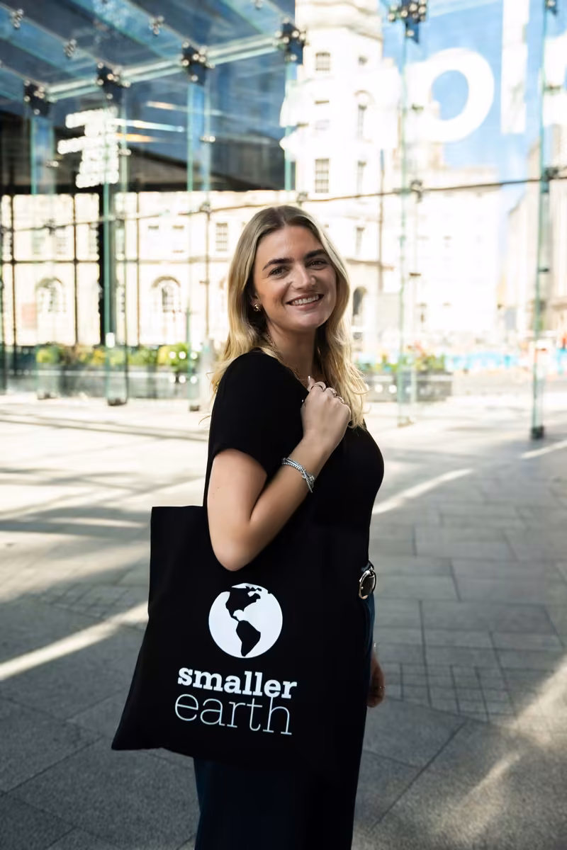 A woman smiling at the camera with a Smaller Earth tote bag over her shoulder, a glass building behind her